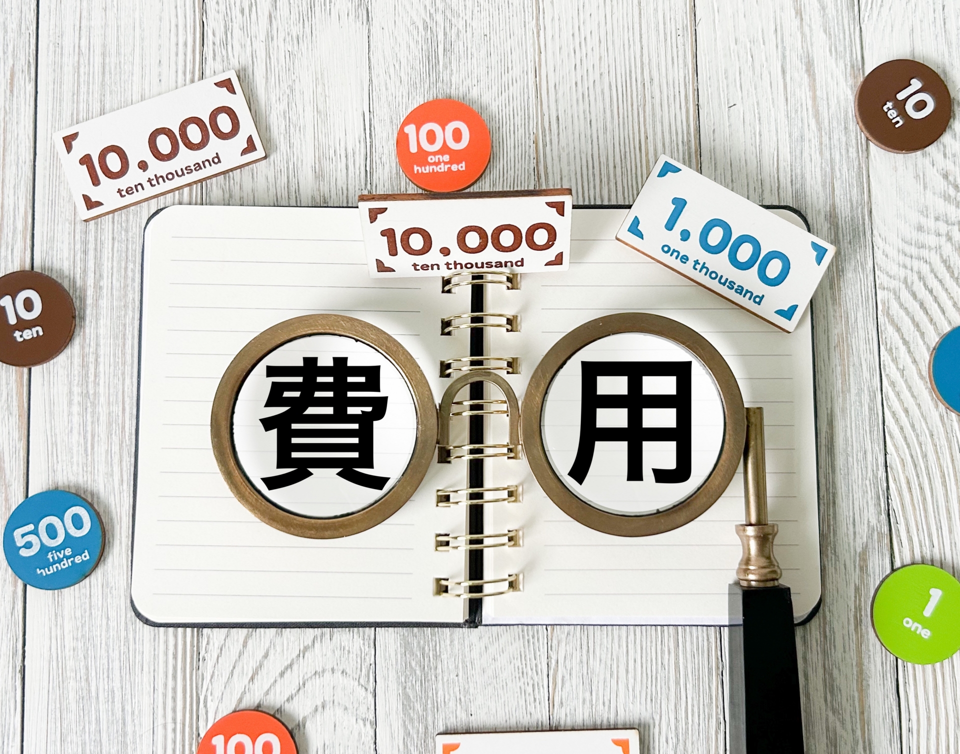 ノートに書かれた「費用」の文字を虫眼鏡で拡大し、周囲に金額チップやカードが置かれている様子