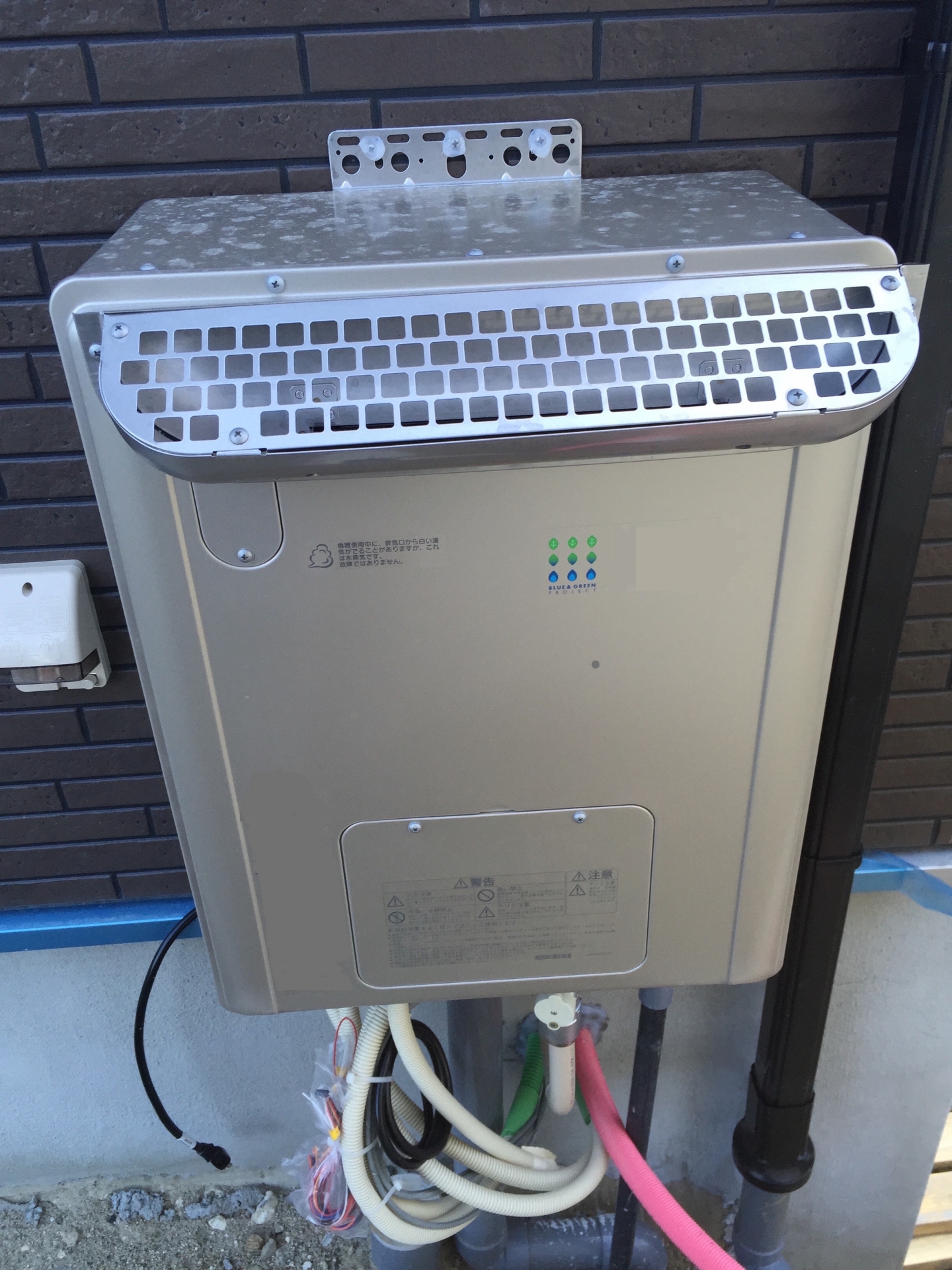 戸建住宅の外壁に並んで設置された古い給湯器と新しい給湯器。配管の接続も確認できる。